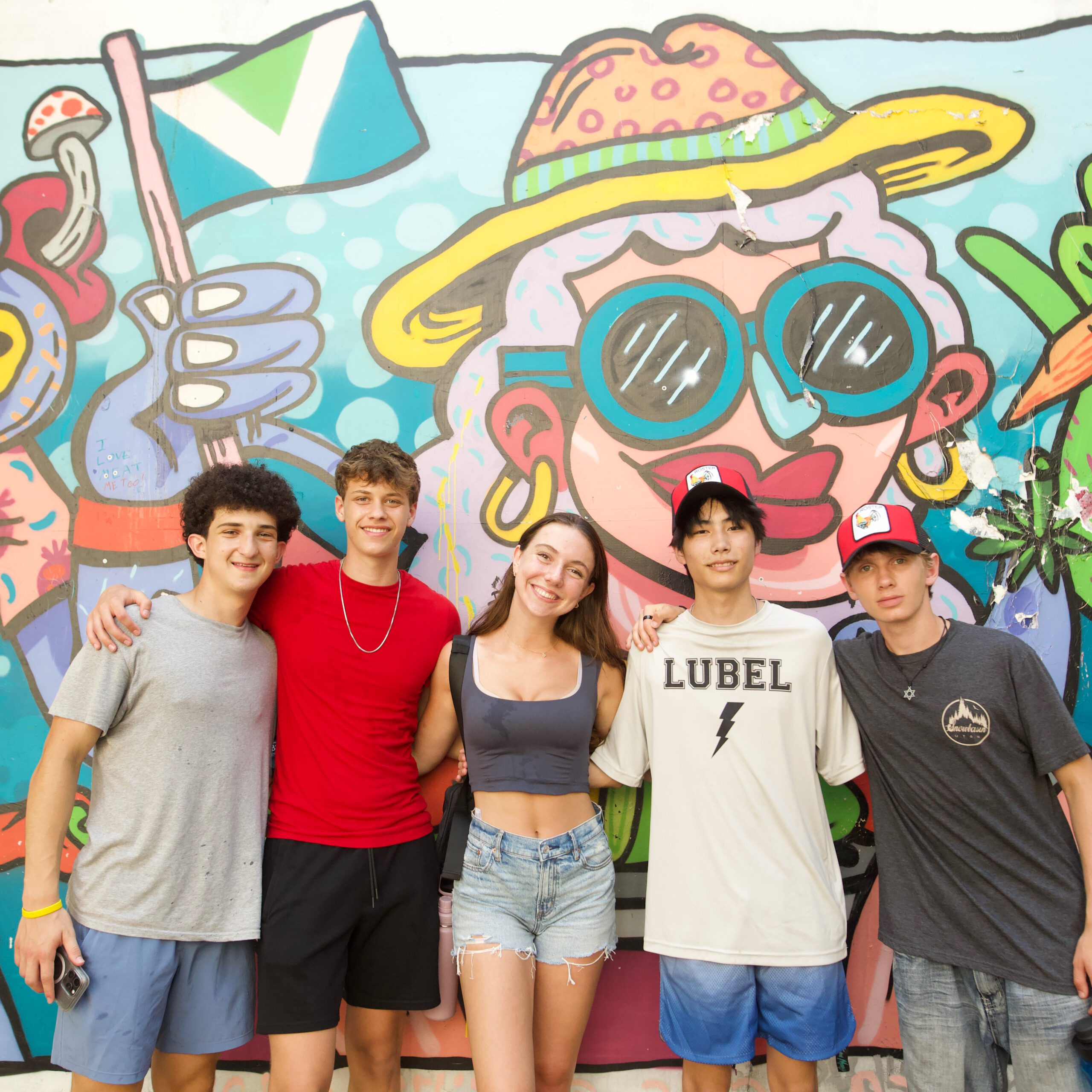ILSI_Header-Photo_Square_A34A1404 Five teens stand arm-in-arm smiling in front of a bright, colorful mural with cartoon-style characters and bold patterns.