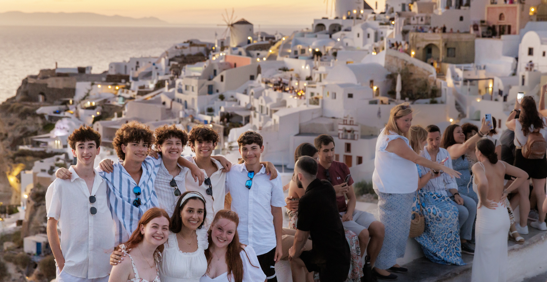 A group of teens poses together at sunset in front of the iconic white buildings and windmills of Santorini, Greece. They are smiling and dressed in light, summery clothing, enjoying the scenic view with other tourists in the background.