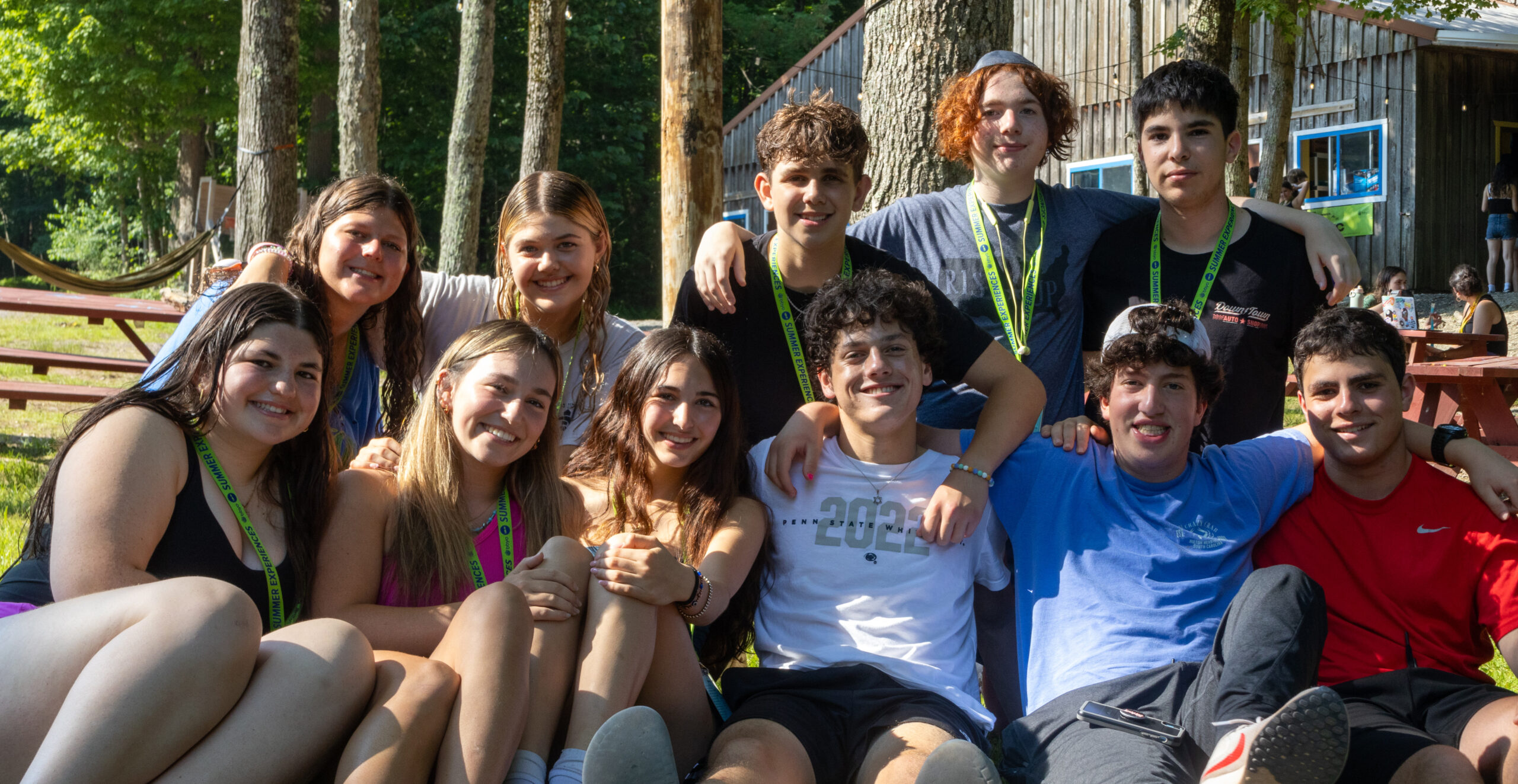 A group of teens sits together outdoors, smiling and relaxing in the sun at a BBYO summer camp. They are wearing BBYO lanyards and enjoying a moment of friendship in a wooded camp setting with picnic tables and hammocks in the background.