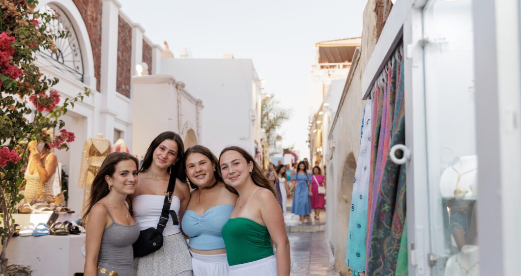Teens standing in an alley in Santorini, Greece. Shops and items for sale line the alley behind them as people bustle through the small area.