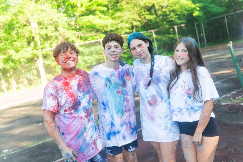 Four teens smile outdoors after a color war activity, their white shirts and faces splattered with bright red, blue, and purple paint.