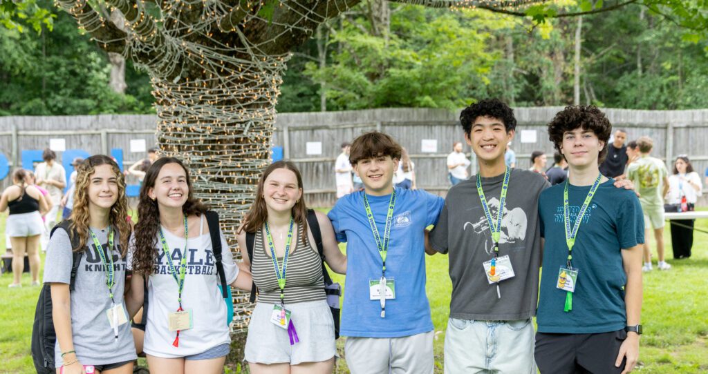 A group of six teens smiling and standing arm-in-arm outdoors in front of a tree wrapped in string lights. They are wearing casual summer clothes and camp-style lanyards with name tags. Other people can be seen mingling in the background on the grass.