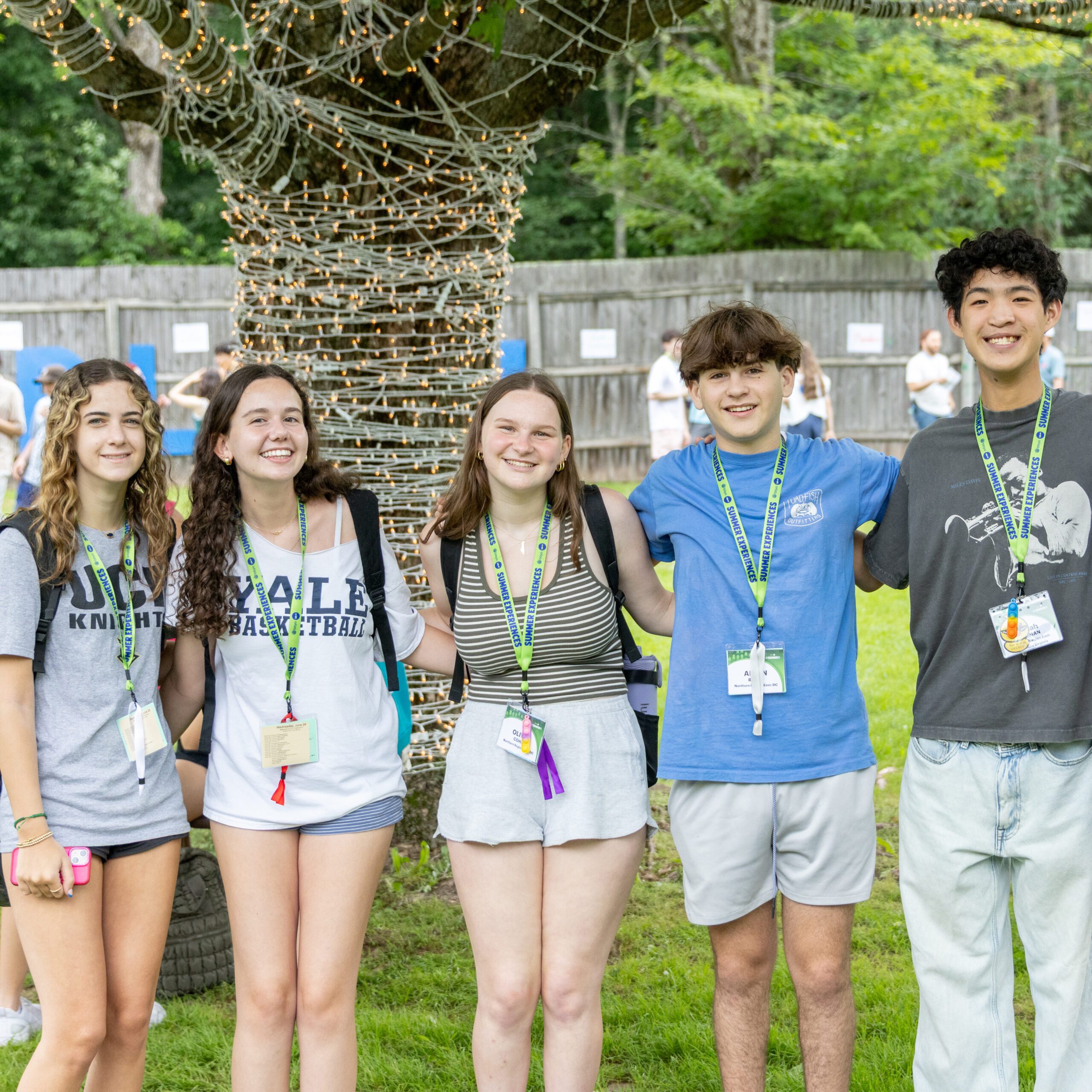 A group of six teens smiling and standing arm-in-arm outdoors in front of a tree wrapped in string lights. They are wearing casual summer clothes and camp-style lanyards with name tags. Other people can be seen mingling in the background on the grass.