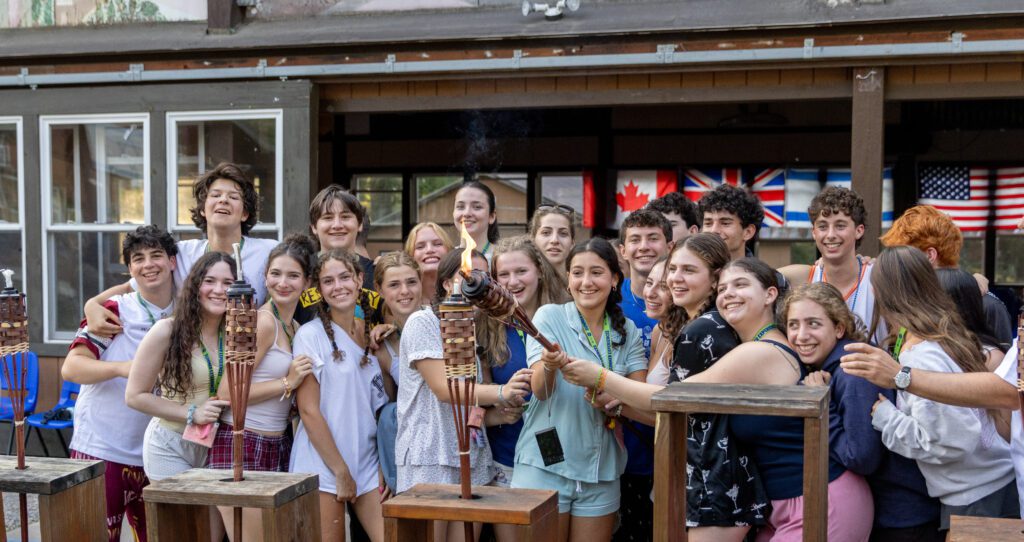 A large group of teens smiling and huddled closely together outdoors, holding a lit ceremonial torch. They are wearing camp-style lanyards and casual clothes, with international flags, including Canadian, British, and American flags, hanging in the background.
