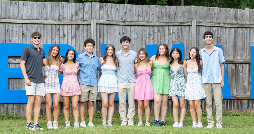 A group of ten teens stands together outdoors, smiling with their arms around each other in front of a wooden fence with large blue letters. They are dressed in summer attire, creating a bright and cheerful atmosphere during a BBYO leadership event.