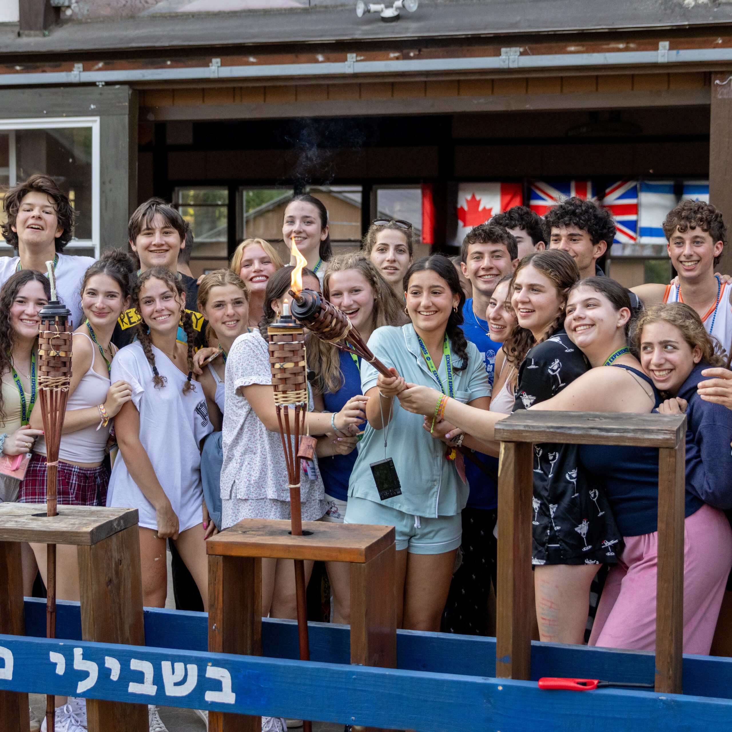 Kallah_Header-Photo_Square_Aaron-Perrotta-ILTC-Jul-04---188 A large group of teens smiling and huddled closely together outdoors, holding a lit ceremonial torch. They are wearing camp-style lanyards and casual clothes, with international flags, including Canadian, British, and American flags, hanging in the background.