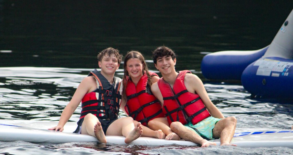 Three teens sitting on a floating board in. the lake at Perlman Camp in Lake Como, PA. The teens have life jackets on and are smiling at the camera.