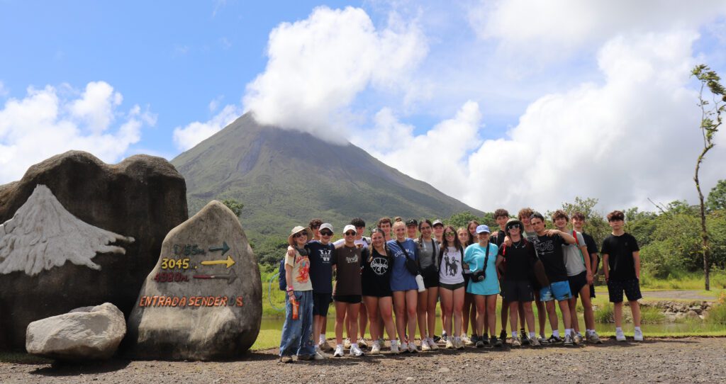 Teens posed for a group photo at the base of Arenal Volcano. It's a partially cloudy day, but you can almost see up to the peak of the volcano.