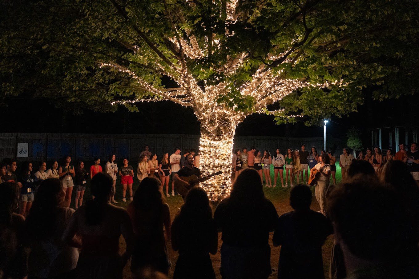 A large group of teens gathered in a circle under a tree adorned with string lights, creating a warm and magical nighttime setting. Two individuals with guitars lead the group in a song or reflection, fostering a sense of community and connection.