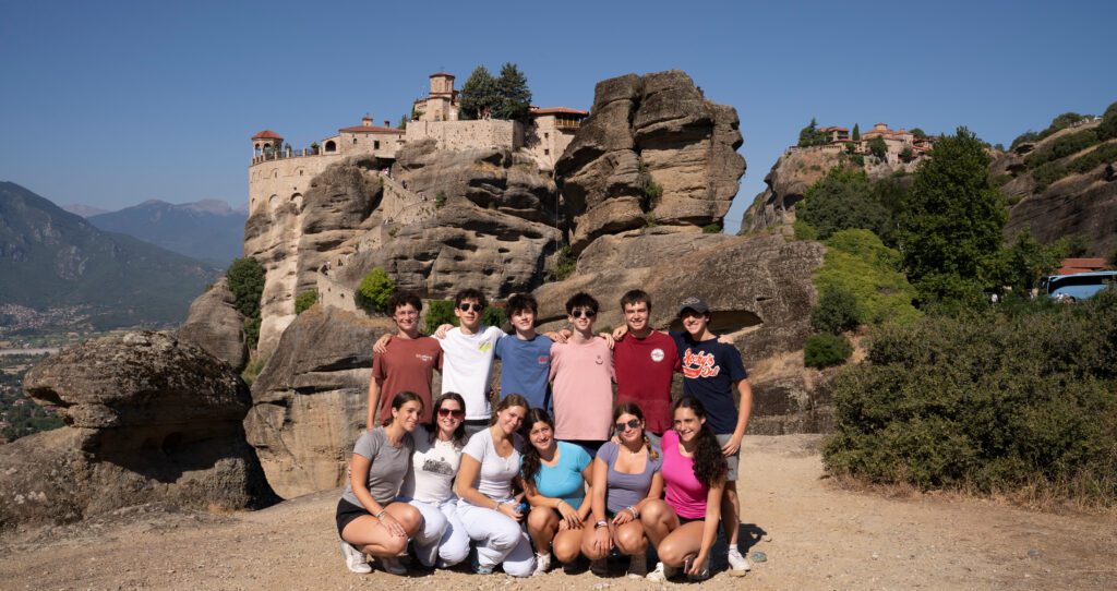Teens posed in front of the Meteor rock formations of Greece. A monastery sits at the top of the rock formation.
