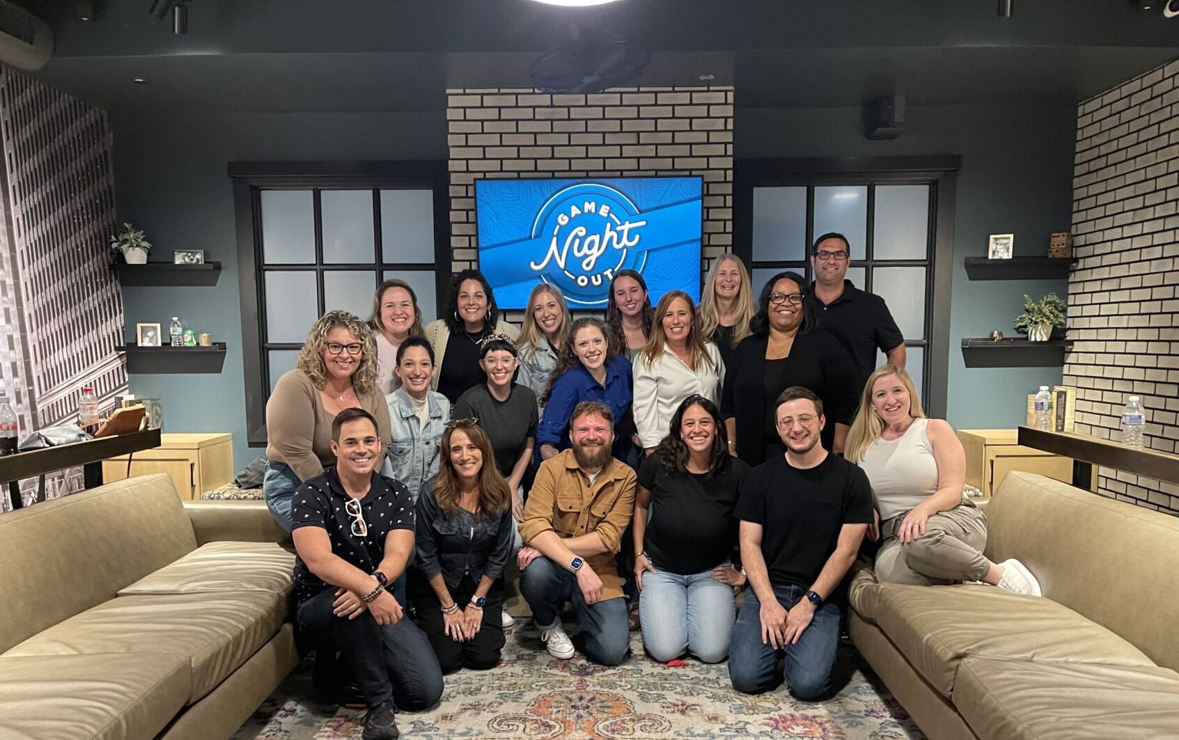 Group of 18 colleagues posing together in a cozy room with a 'Game Night Out' sign in the background. They are smiling and gathered in front of a brick wall and couches, creating a friendly and relaxed atmosphere.