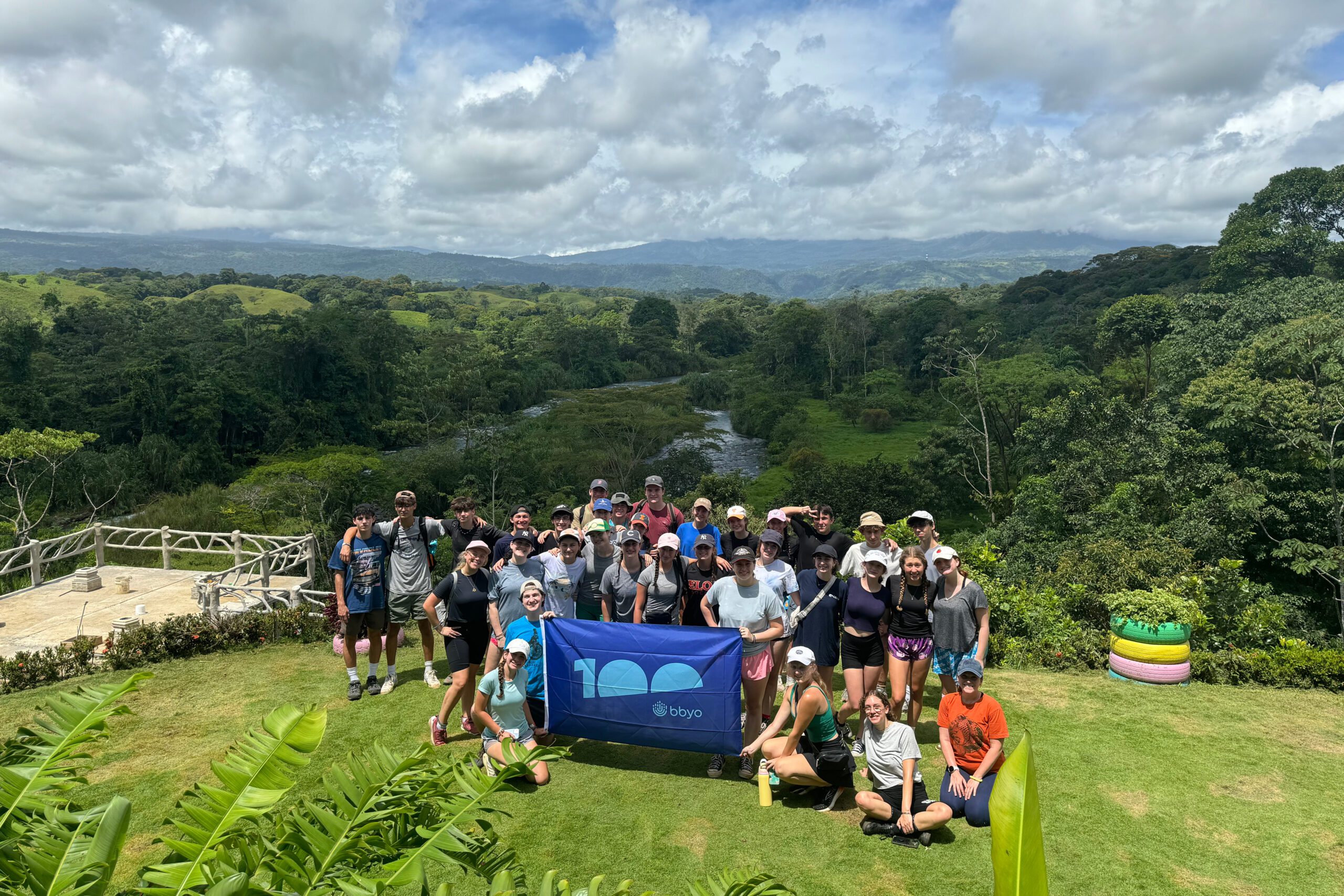 A large group of teens poses together on a lush green hilltop, holding a blue BBYO 100th anniversary flag. Behind them, a river winds through a dense tropical forest, with mountains visible in the distance under a partly cloudy sky. The group is dressed in outdoor gear, including hats and backpacks, suggesting they are on a hiking adventure. The scene captures the beauty of nature and the camaraderie of a group expedition.