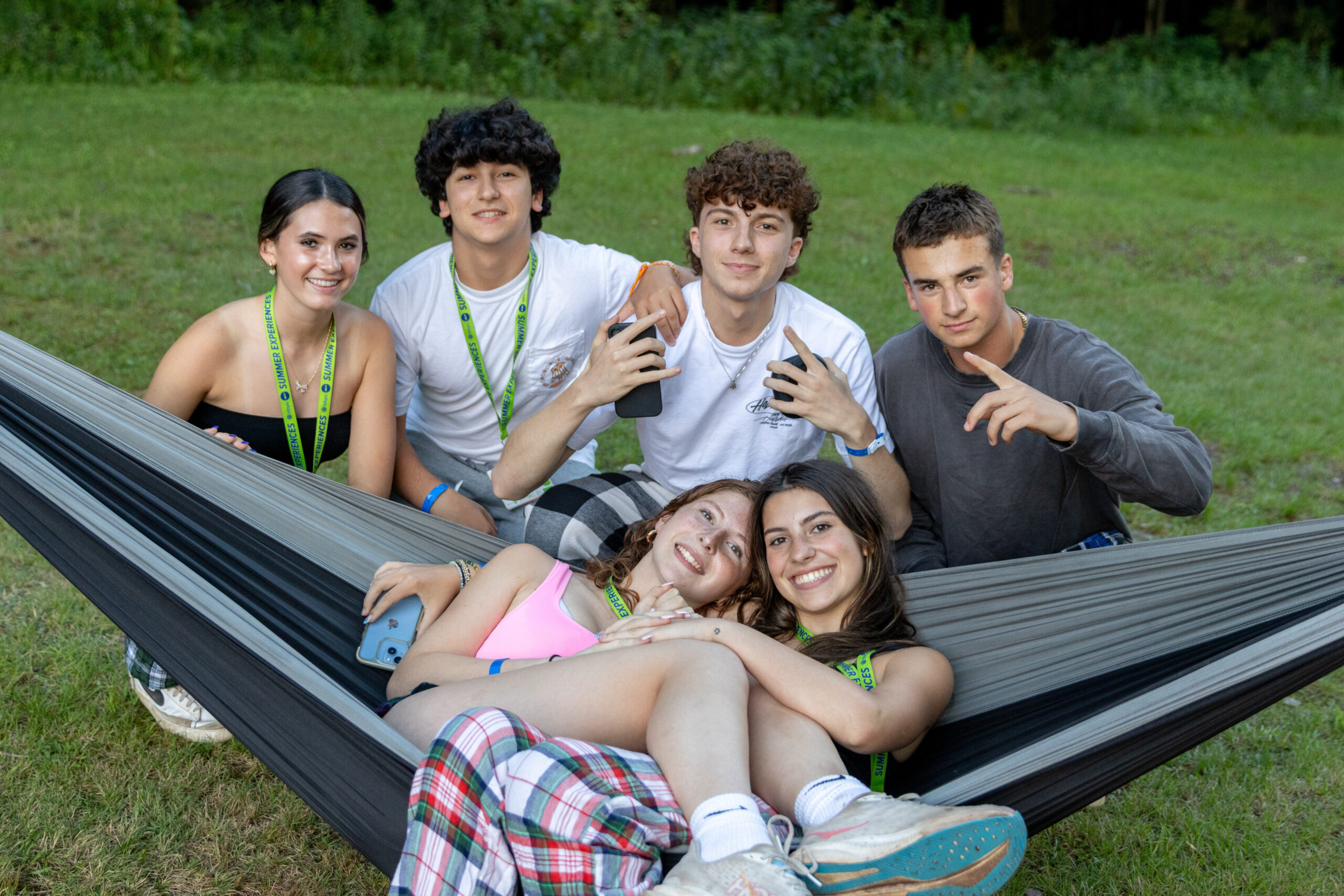 A group of six teens relaxes together outdoors, with two lying in a hammock and the others sitting nearby, sharing a moment of fun and relaxation during a BBYO summer camp program.