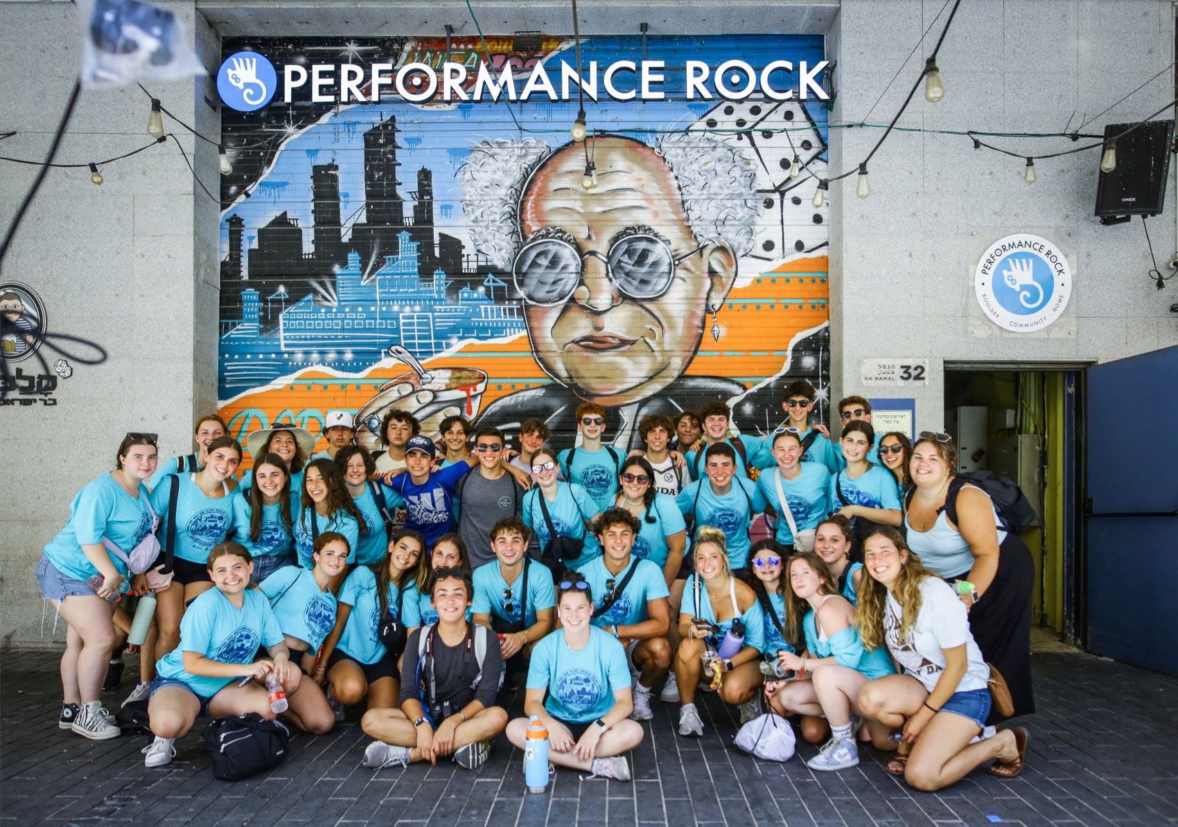 A large group of teens pose together in front of a colorful mural at 'Performance Rock,' depicting an iconic figure in sunglasses with a cityscape in the background. The group is smiling, wearing matching blue shirts, and enjoying their BBYO summer leadership experience.