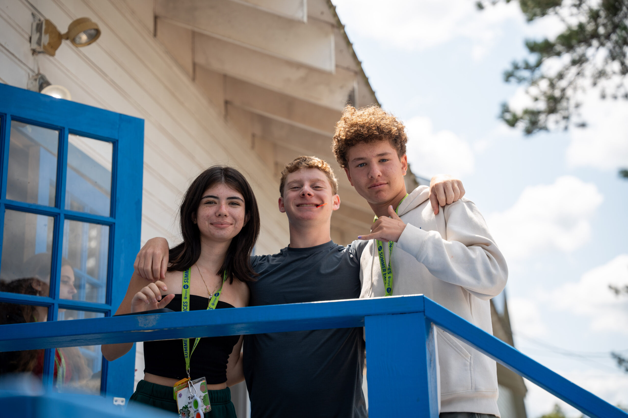 Three teens stand on a porch, arm-in-arm, smiling and enjoying a sunny day together at a summer leadership camp program.