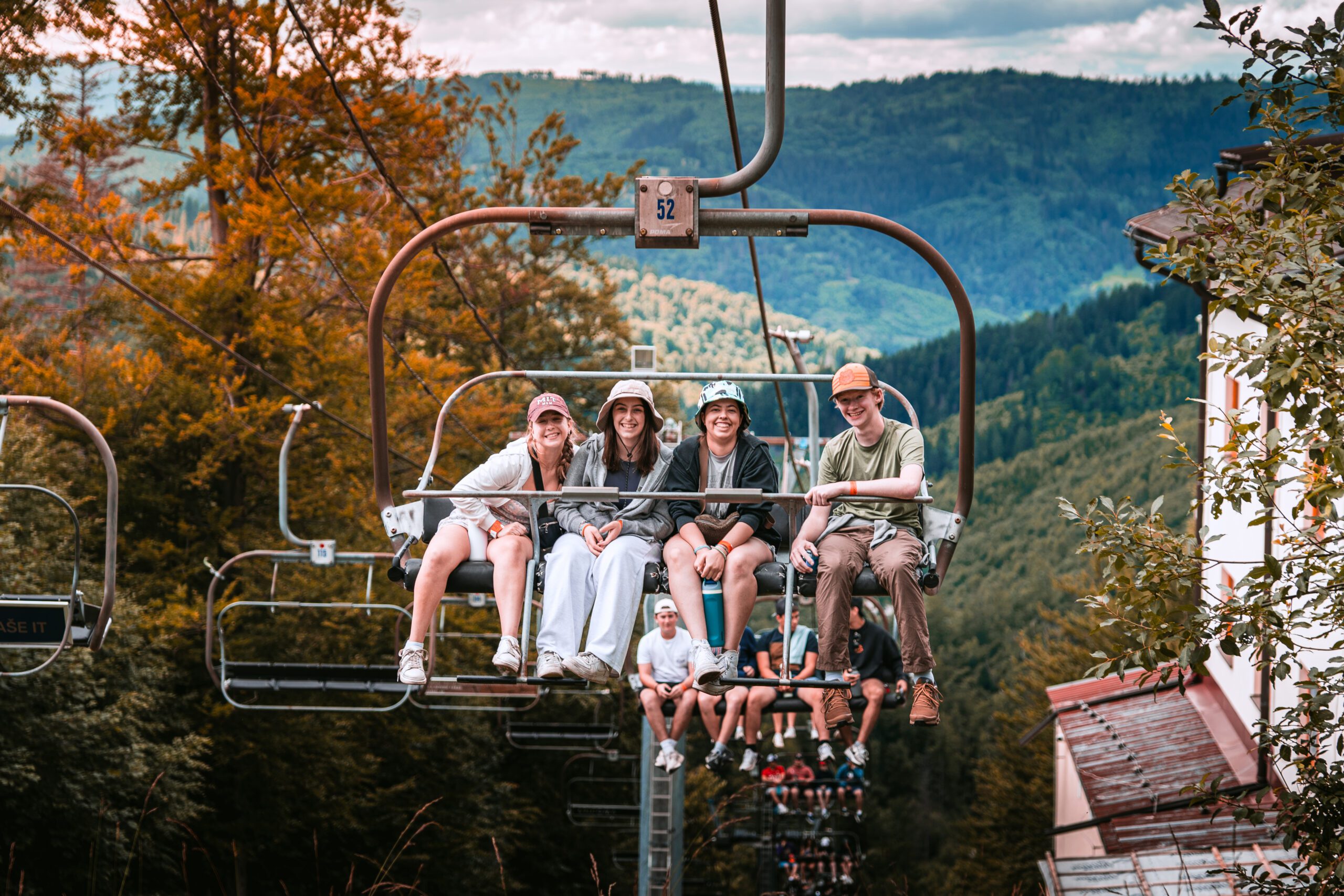 A group of teens smiles and laughs while sitting on a ski lift, enjoying the scenic mountain views around them during their BBYO adventure.