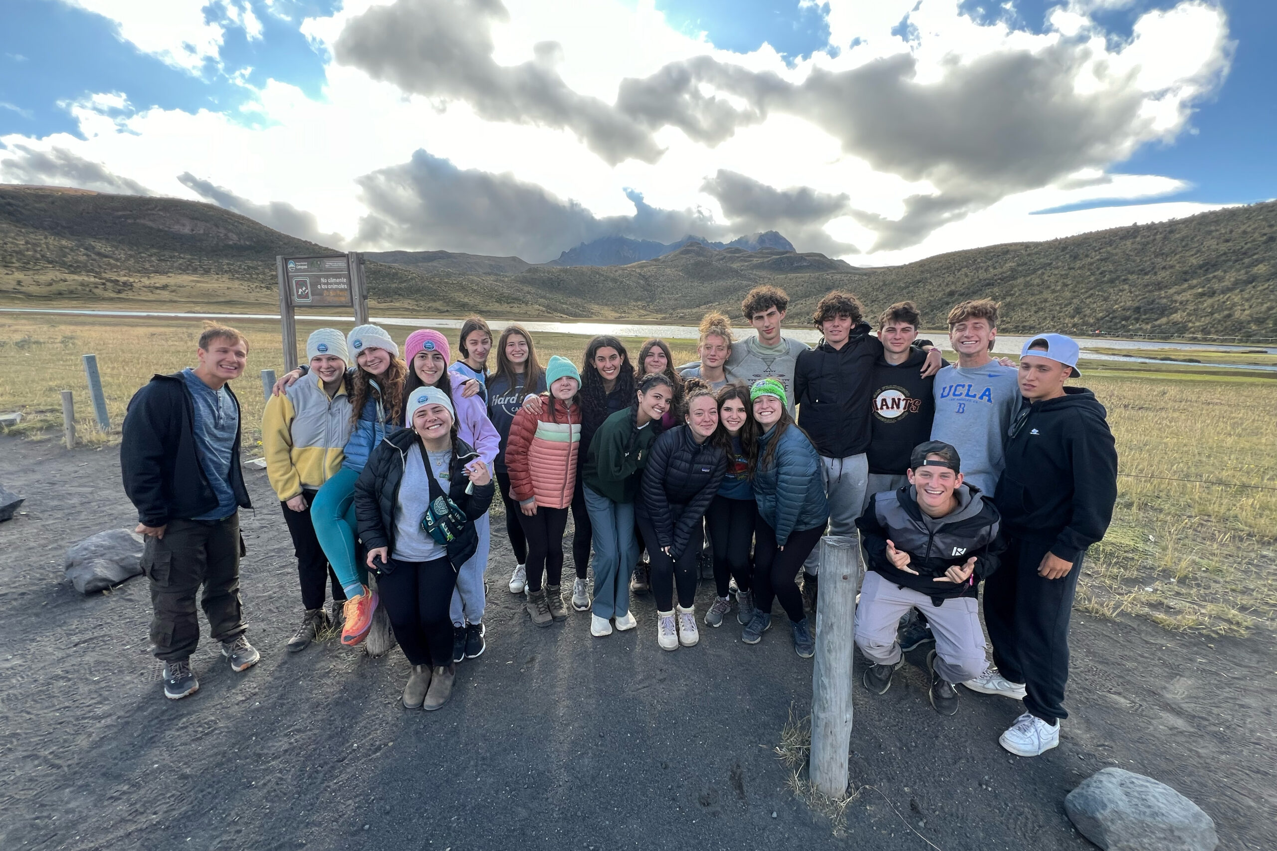 A group of teens bundled up in jackets and hats poses together outdoors with a dramatic mountain backdrop during a BBYO hiking adventure.