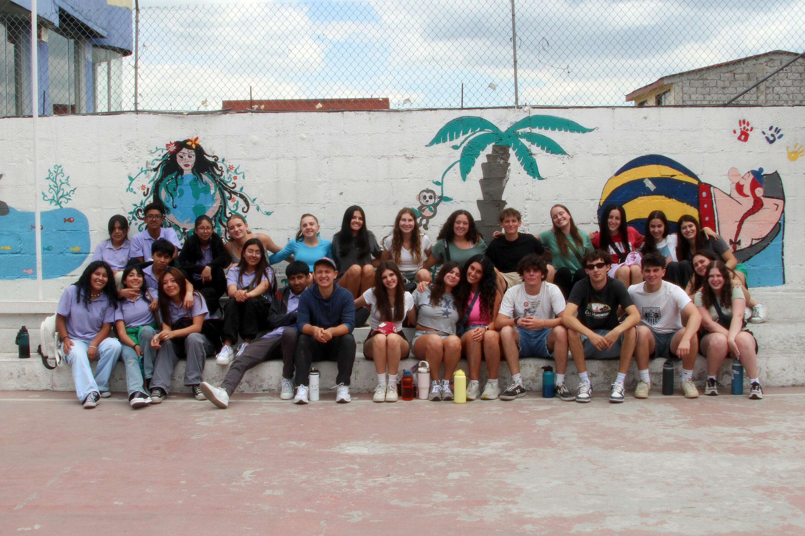 A large group of teens sit together in front of a colorful mural in an outdoor setting, enjoying a moment of togetherness during their BBYO summer program.