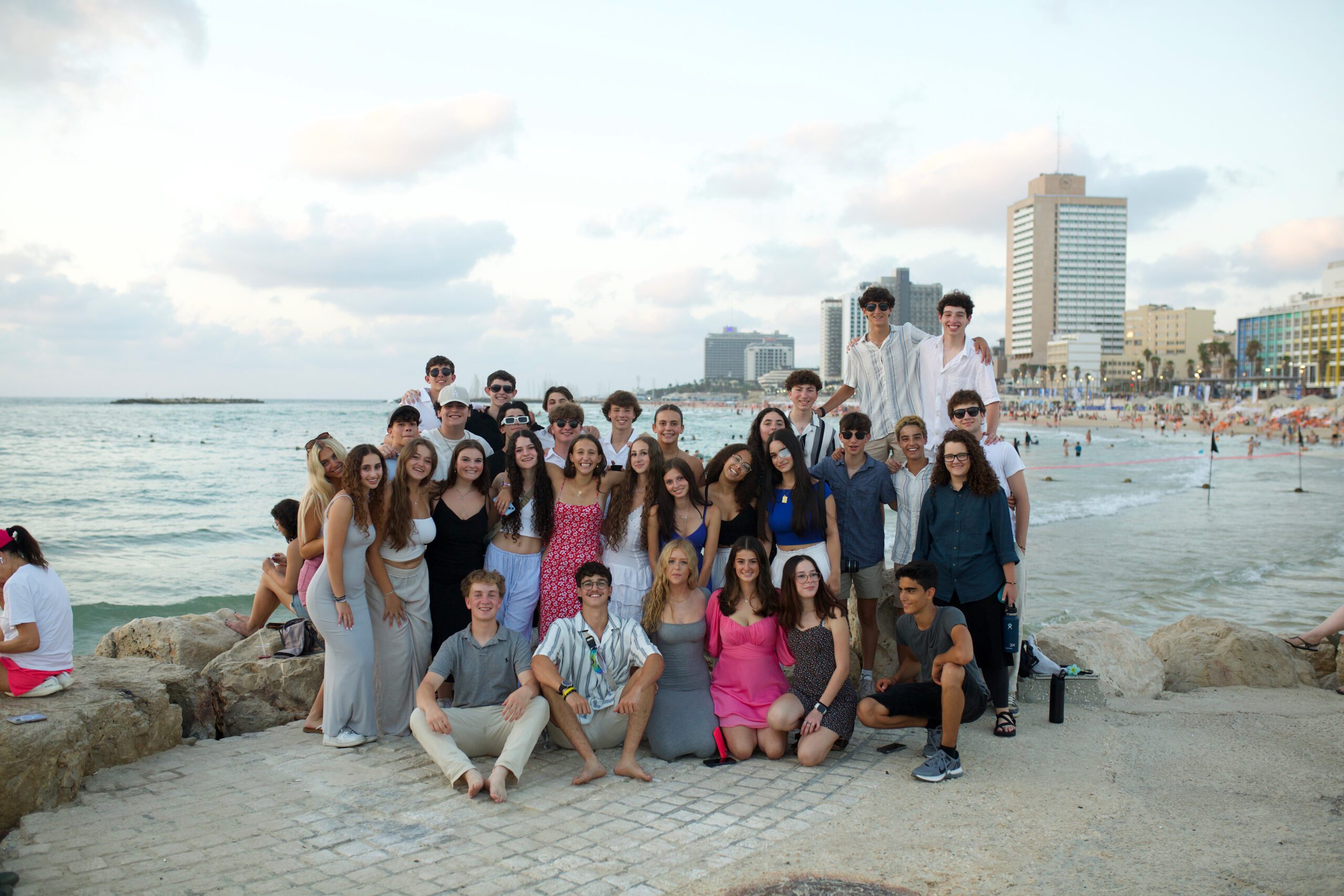 A large group of teens poses together on a beachfront promenade with the sea and city skyline in the background. They are dressed in casual summer clothes, enjoying a sunset moment during their BBYO leadership experience in Tel Aviv.