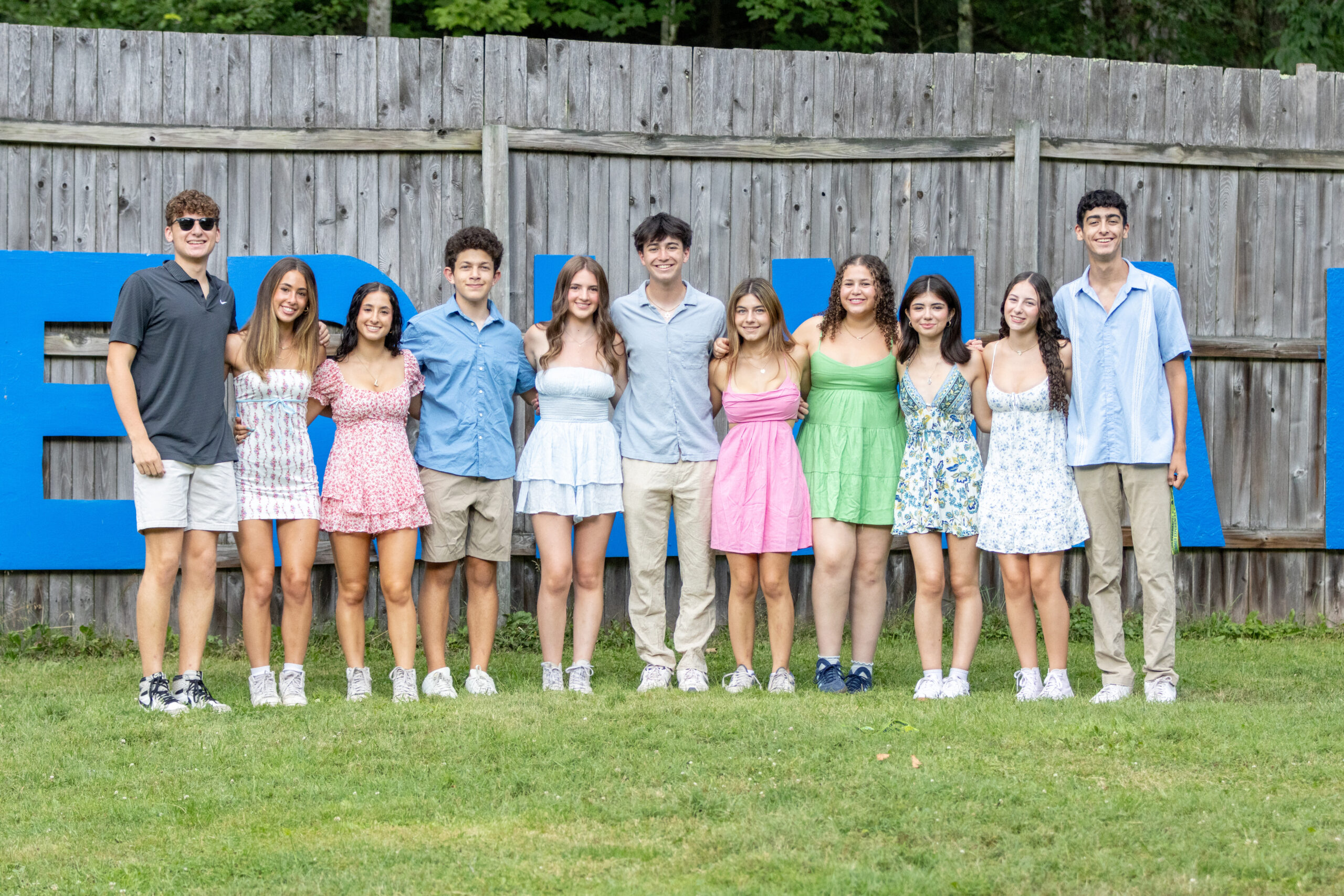 A group of ten teens stands together outdoors, smiling with their arms around each other in front of a wooden fence with large blue letters. They are dressed in summer attire, creating a bright and cheerful atmosphere during a BBYO leadership event.