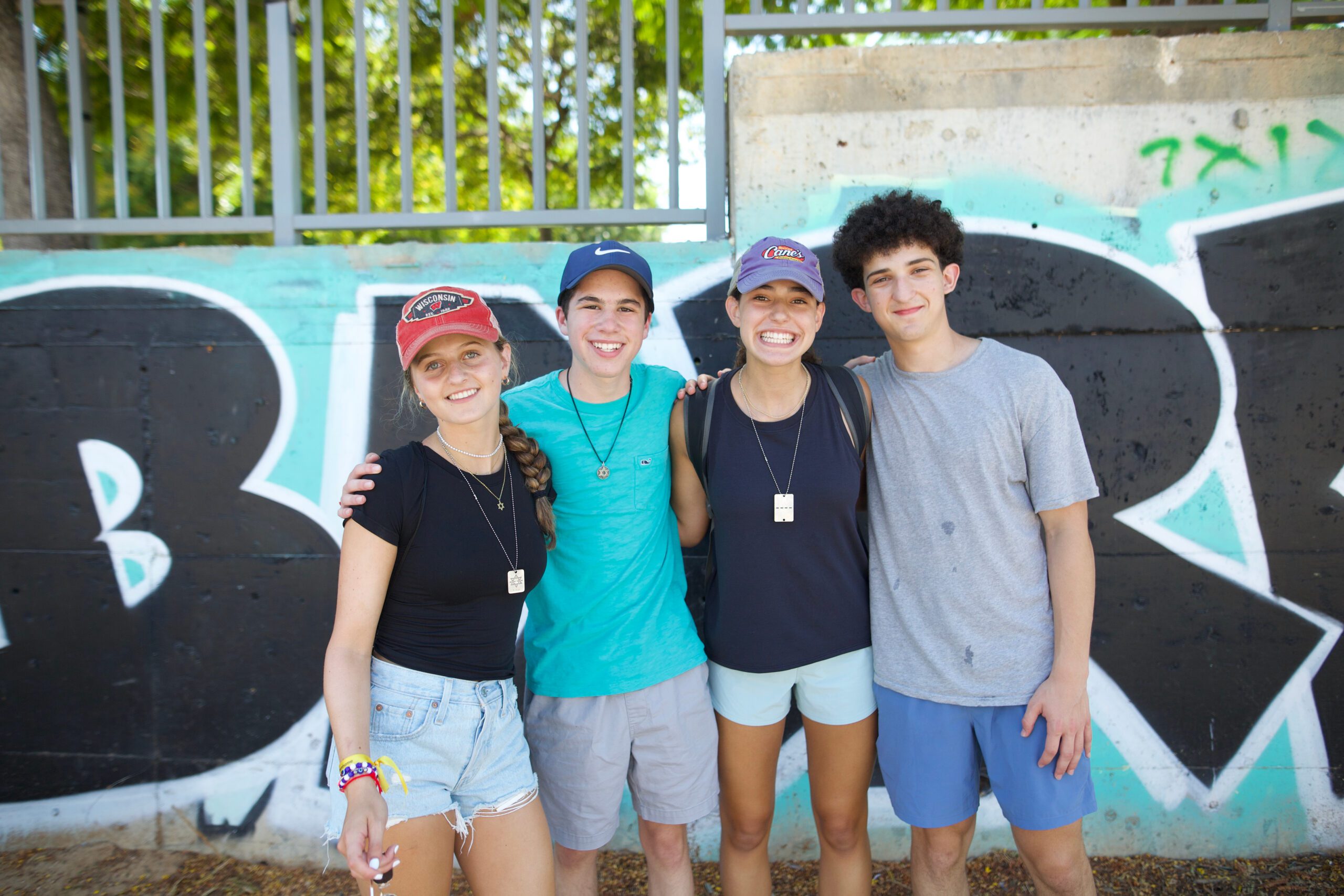 Four teens stand together smiling in front of a colorful graffiti wall, wearing casual clothes and hats, enjoying a fun moment during their BBYO summer experience.