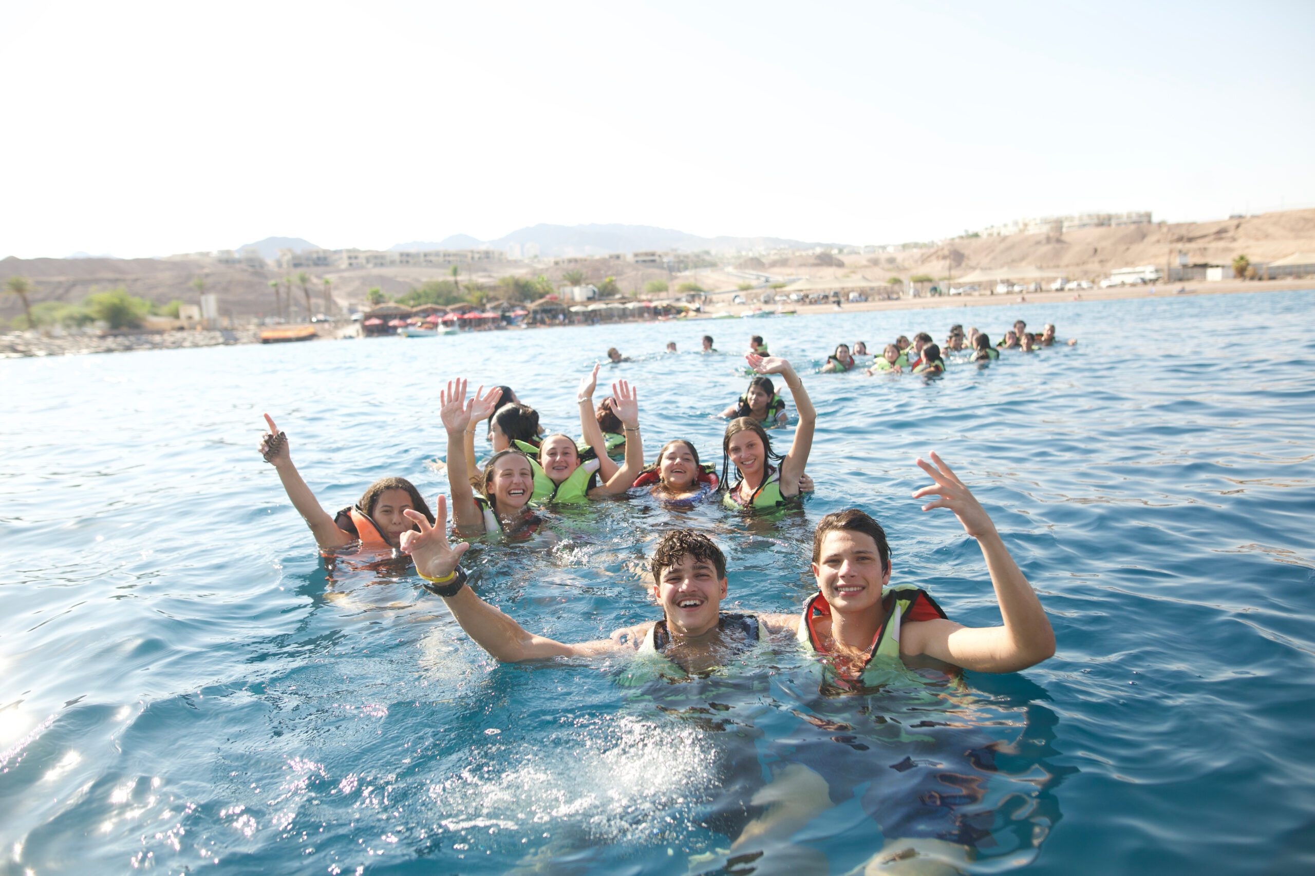 A group of teens in life vests happily swimming together in open water, waving and smiling during a BBYO summer program activity.