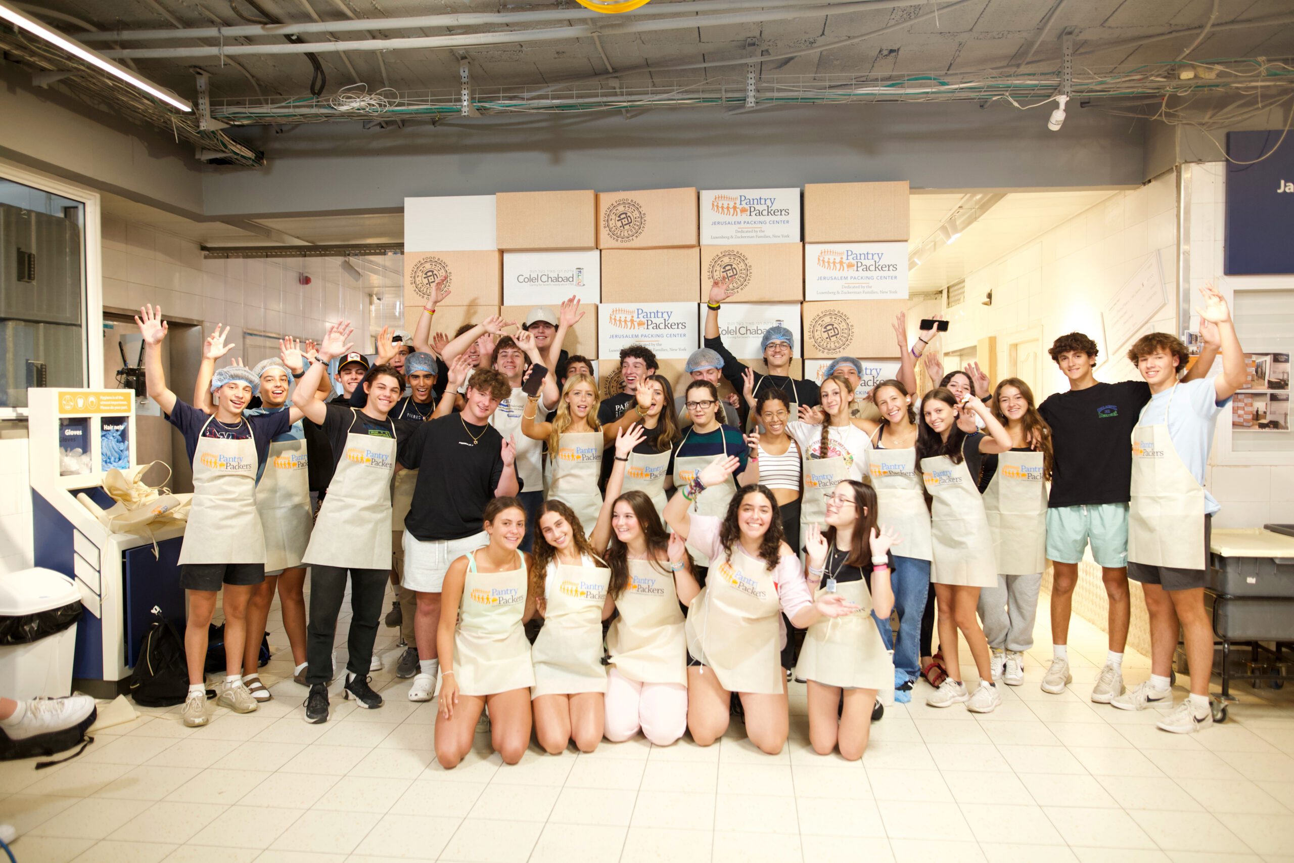 A large group of teens wearing aprons and gloves stands together inside a volunteer kitchen, raising their hands and smiling as they participate in a community service activity.