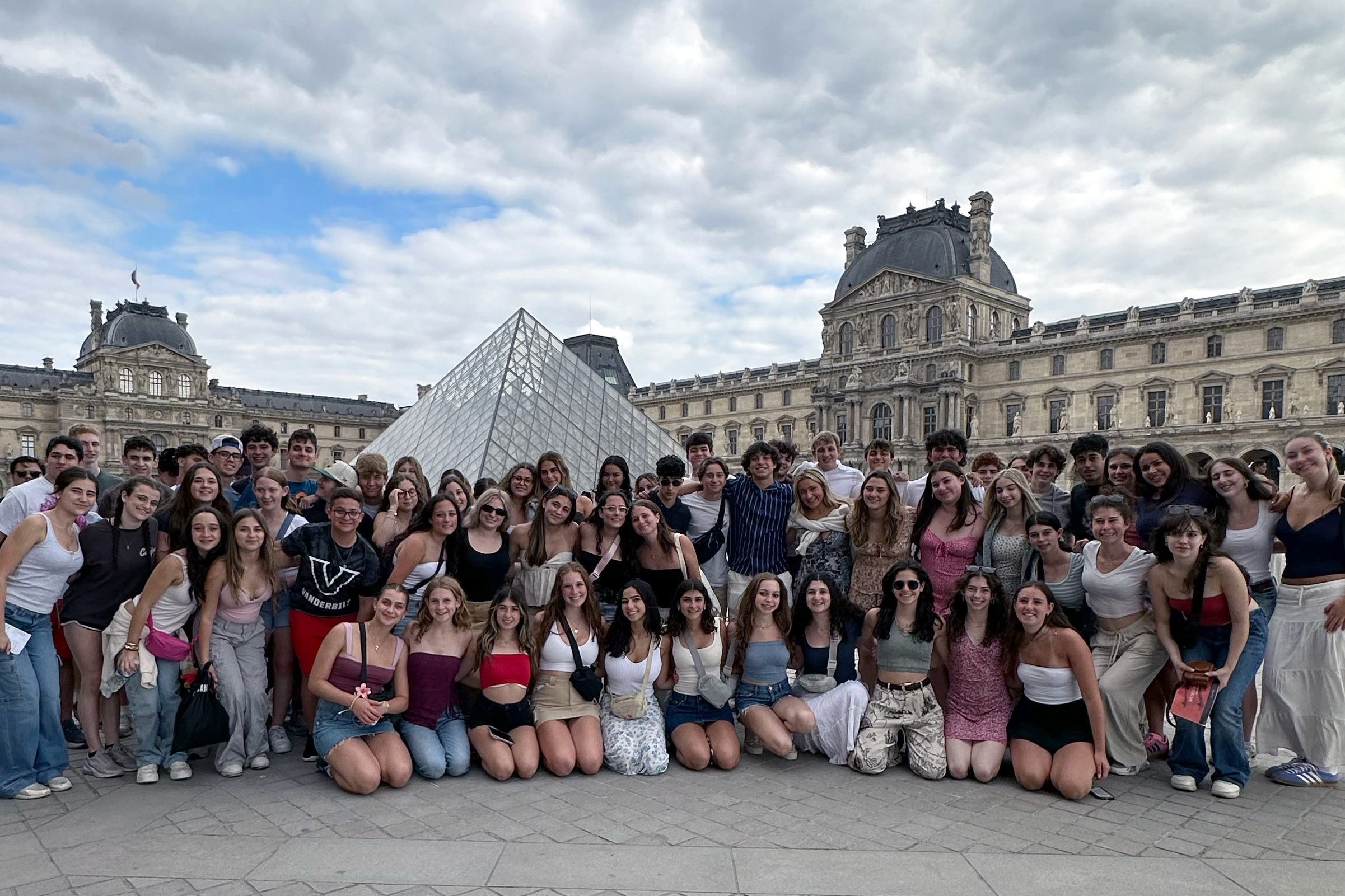 A large group of teens smiling and posing for a photo in front of the glass pyramid entrance to the Louvre Museum in Paris, with the historic Louvre Palace visible in the background under a partly cloudy sky.