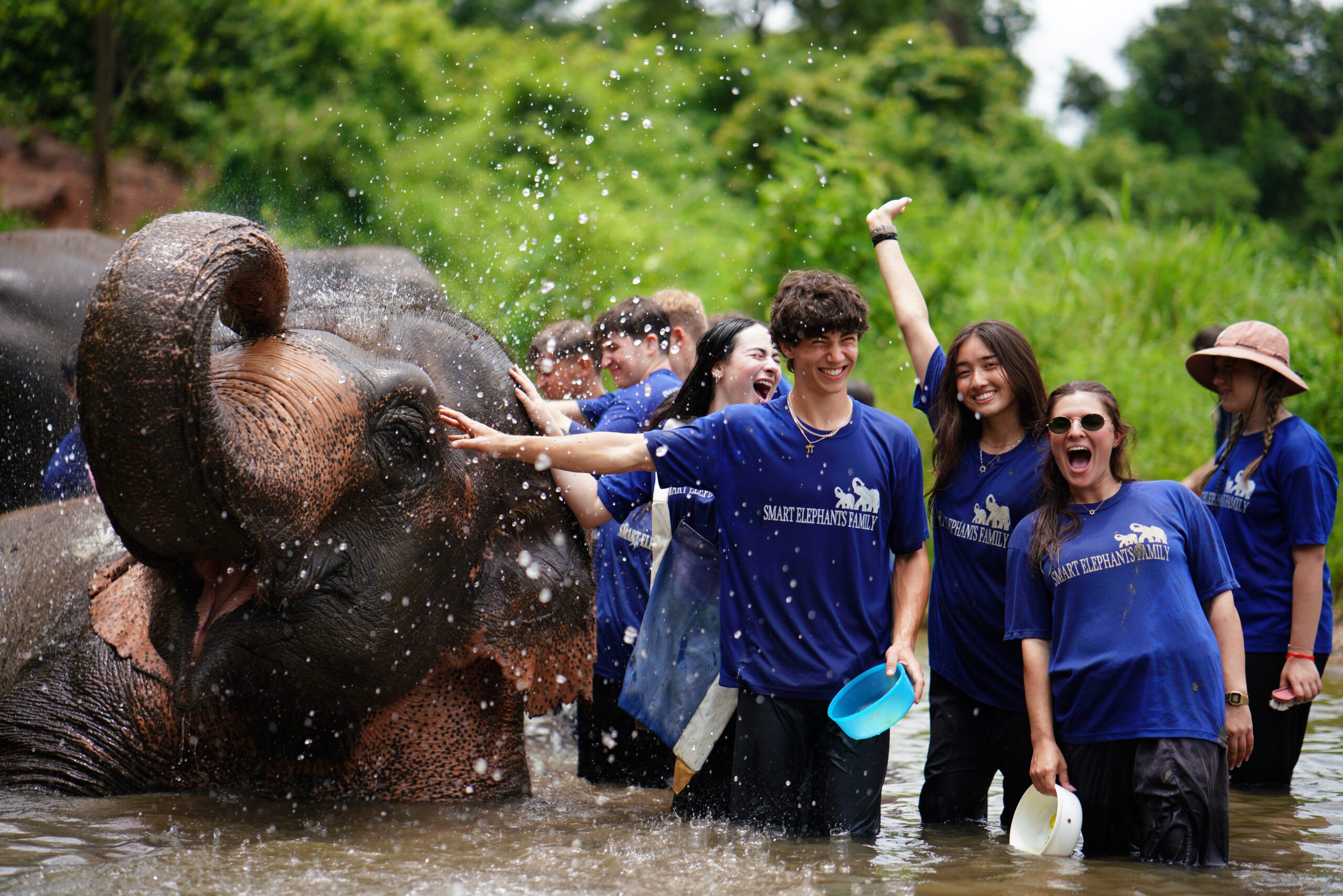 A group of smiling teens wearing matching blue "Smart Elephants Family" t-shirts playfully bathe elephants in a shallow river. Water splashes around them as they interact with the elephants, creating a joyful and energetic scene surrounded by lush greenery.