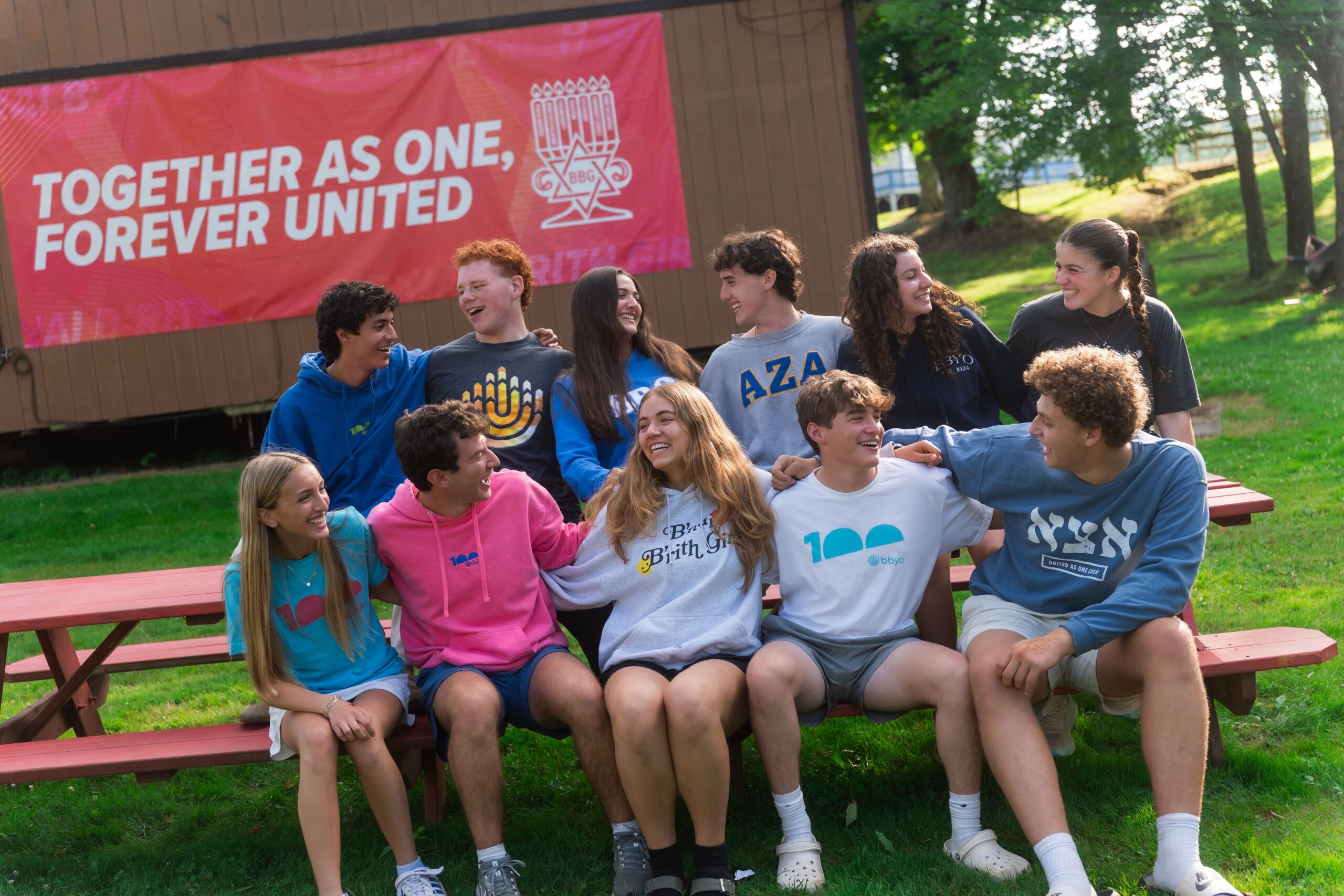 A group of smiling teens sit together on a red picnic bench, laughing and leaning into each other, with a large red banner behind them reading “TOGETHER AS ONE, FOREVER UNITED.”