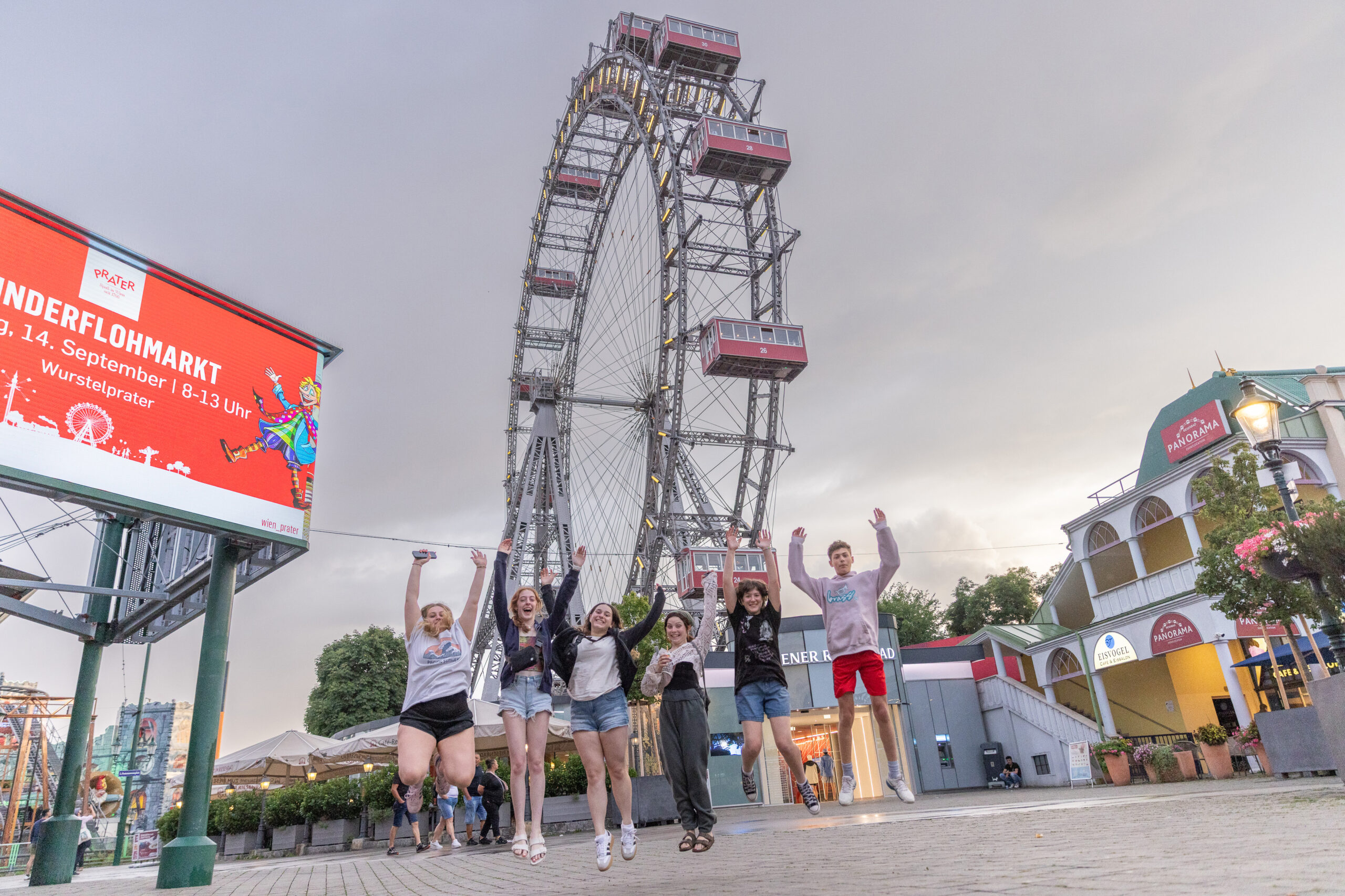 A group of six teens jump with their hands in the air in front of Vienna’s iconic Prater Ferris wheel, smiling and enjoying the amusement park atmosphere.