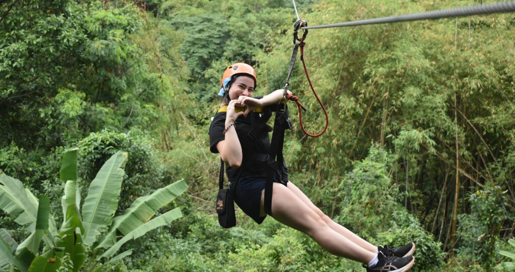 A teen wearing a helmet and harness smiles while ziplining through a lush green forest, making a heart shape with their hands.
