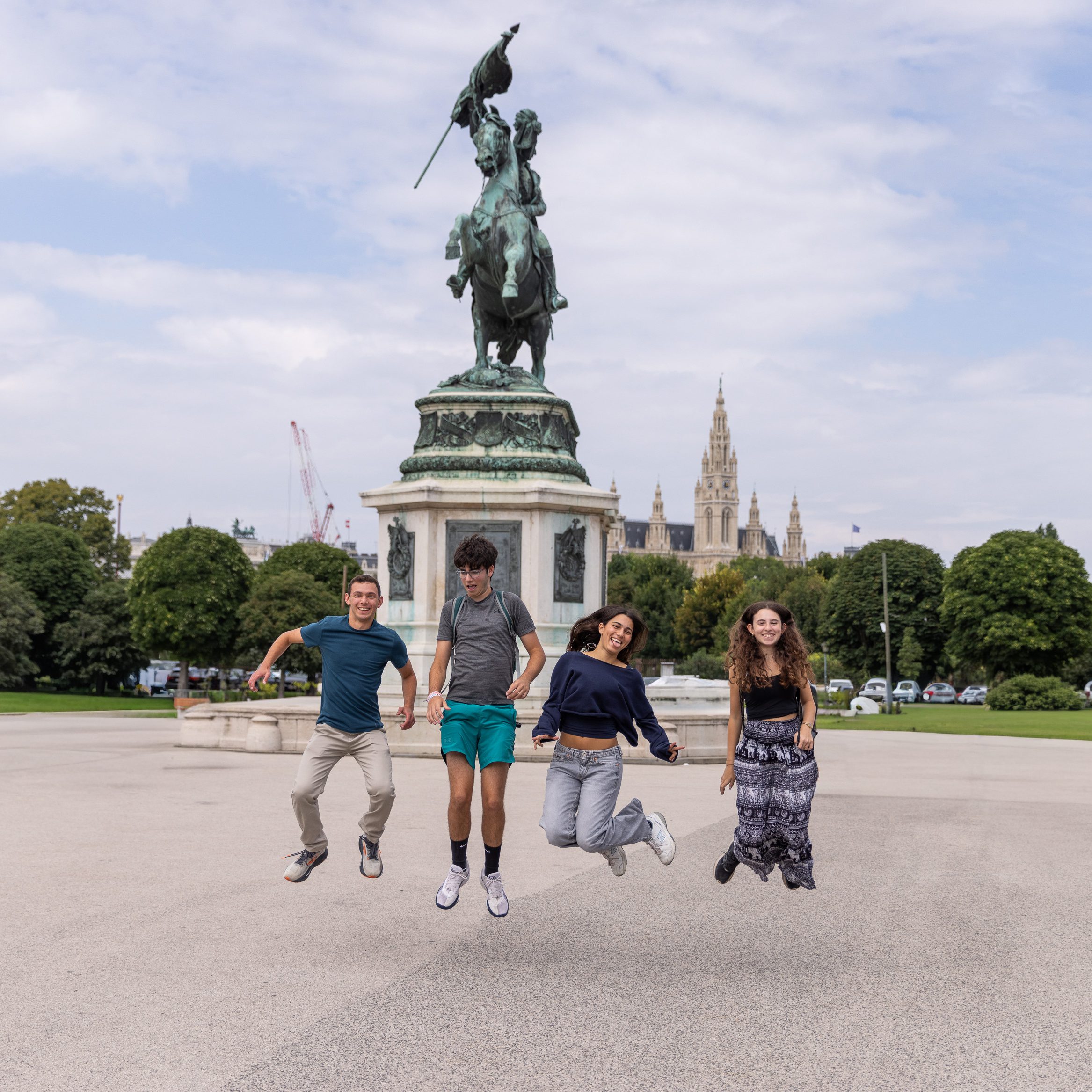 Central-Europe-Discovery_Header-Photo_Square_2Q9A1476 Four teens jump in the air smiling in front of a large equestrian statue in Vienna, Austria, with the spires of the Rathaus in the background and groups of people walking nearby.