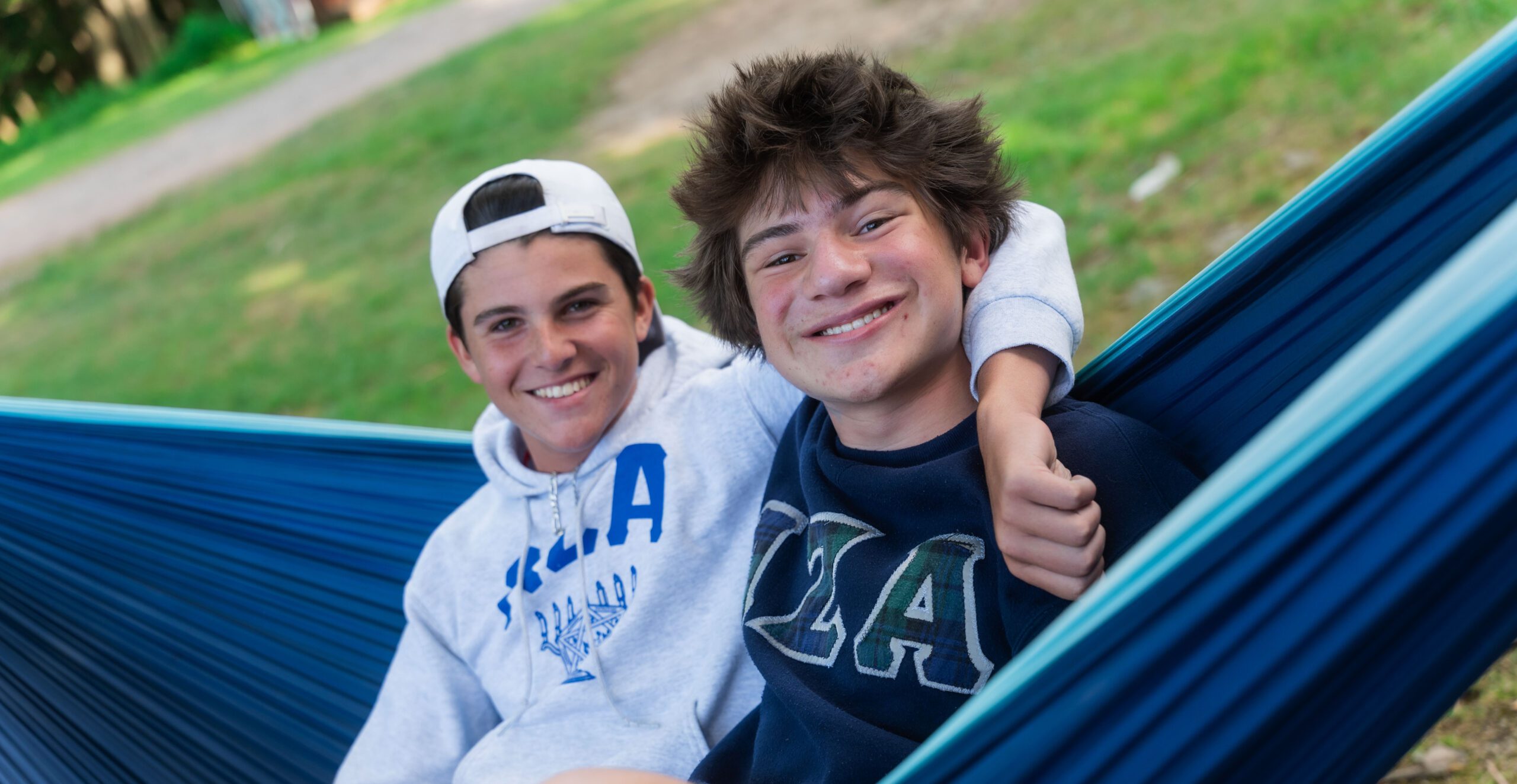 FY26_Our-Programs_Camp_CLTCPhotoshoot2025-1015760 Two teens relax in a blue hammock outdoors, smiling and wearing BBYO sweatshirts while enjoying a peaceful camp day.