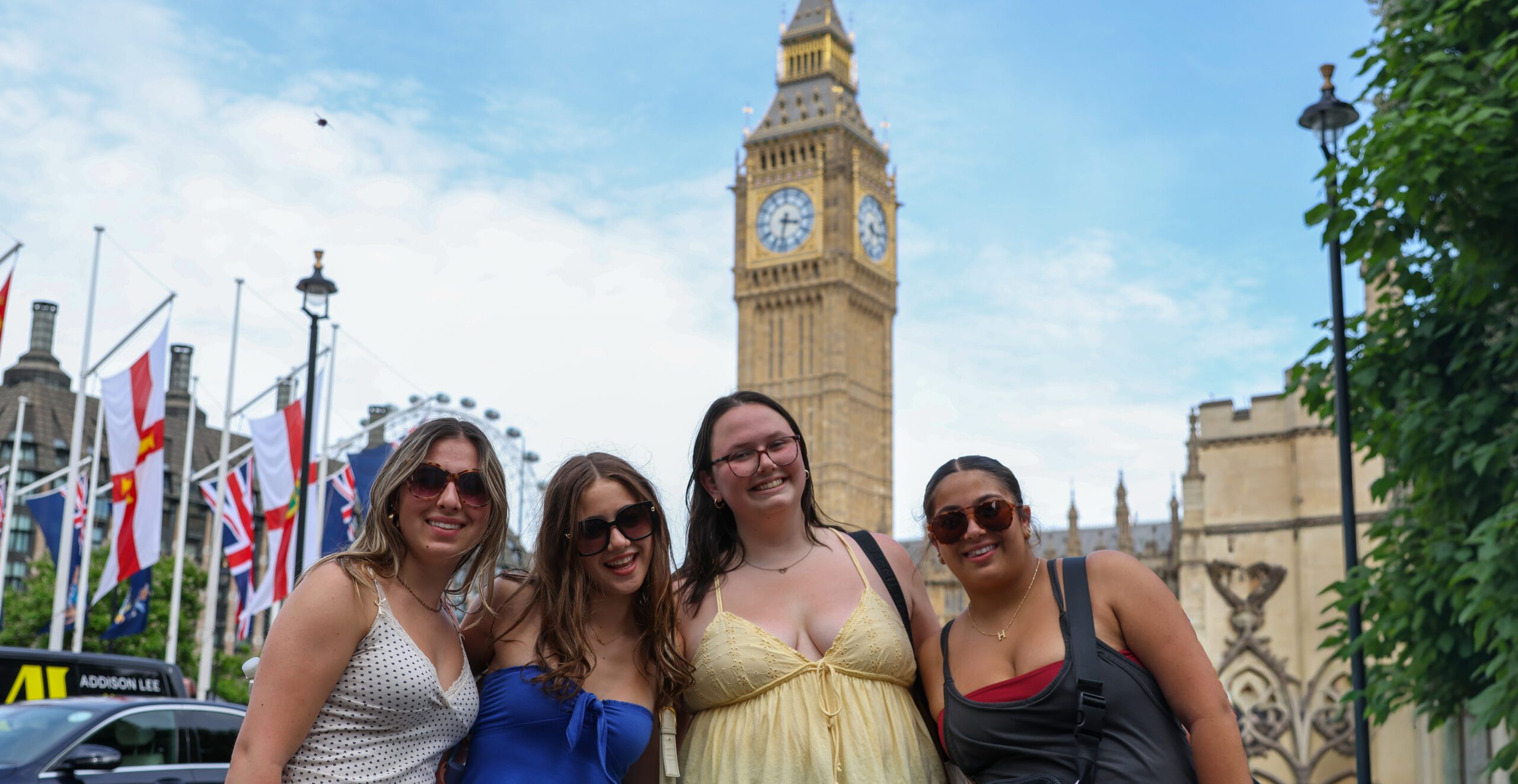 FY26_Our-Programs_PassportBBYO-Passport-day-2-26 Four teens smile together in front of Big Ben in London, posing with flags and city landmarks in the background on a sunny day.