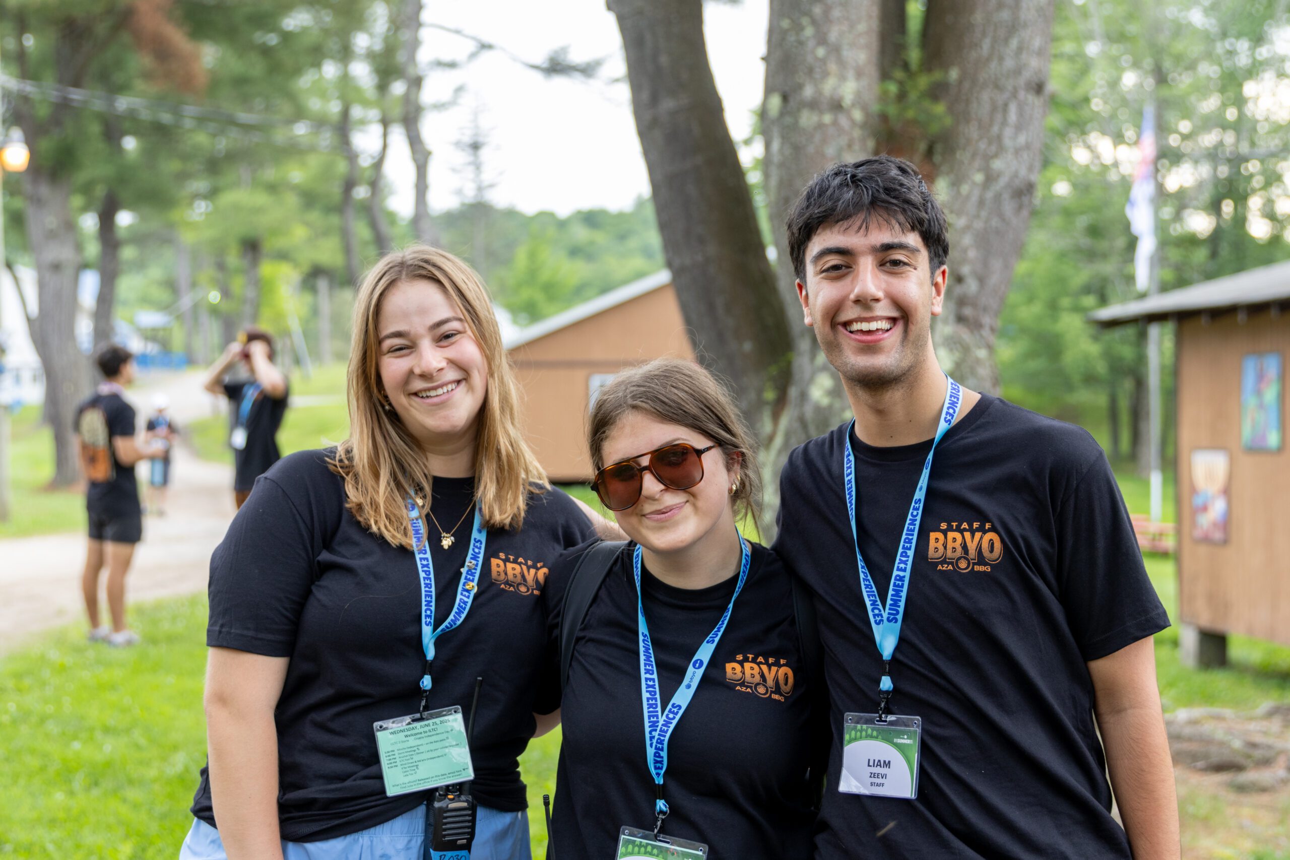 Three BBYO staff members smile together outdoors at camp, wearing black staff shirts and blue lanyards.