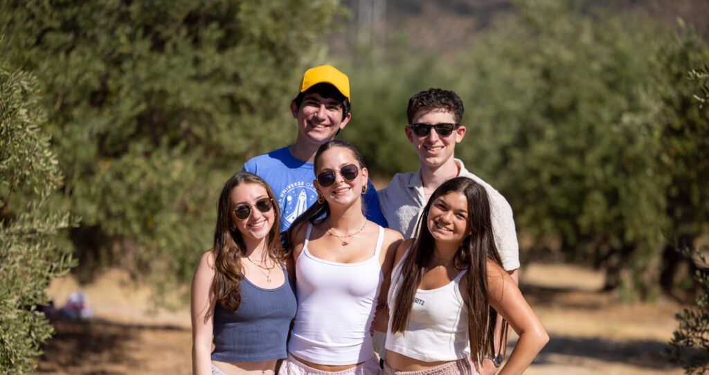 Five teens smile and pose closely together in a sunny olive grove, wearing casual summer outfits and sunglasses.