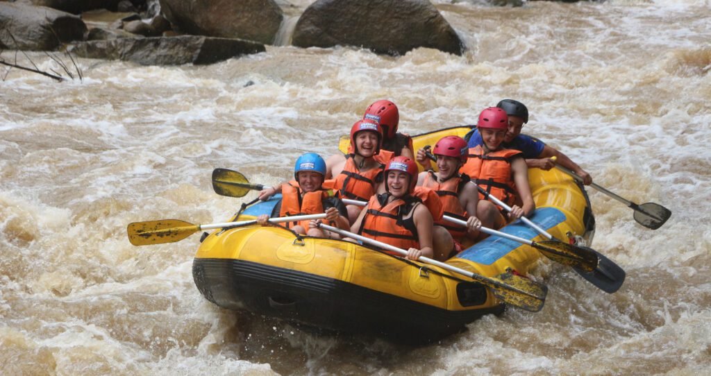 A group of teens wearing helmets and life jackets paddle together in a yellow raft, smiling as they navigate rushing whitewater rapids.
