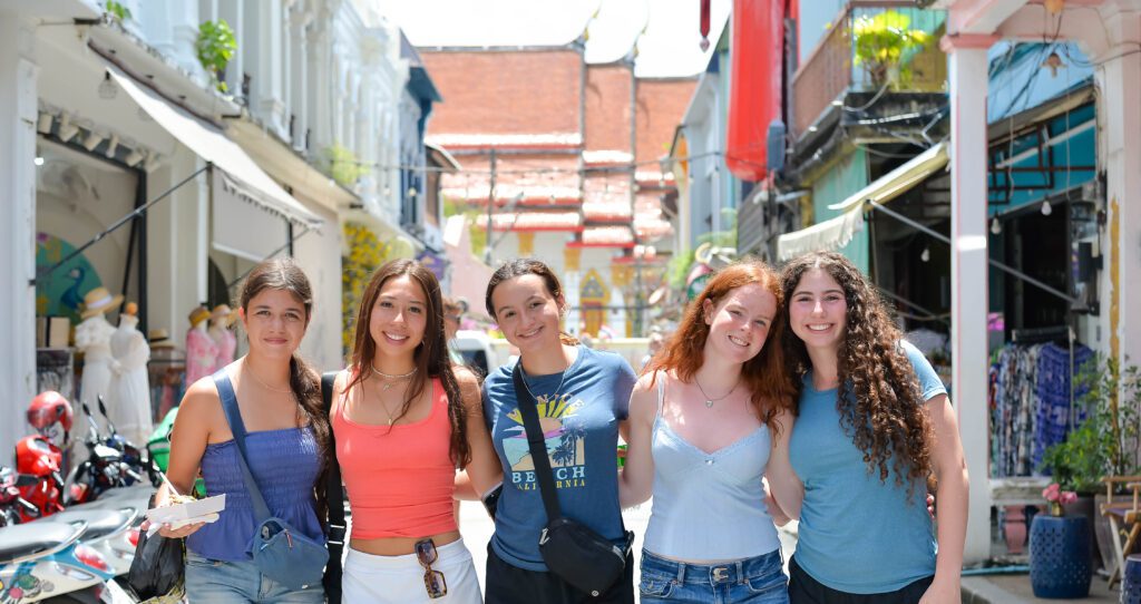 Five teens stand arm-in-arm smiling on a colorful street with shops and motorbikes, with a traditional temple building visible in the background.