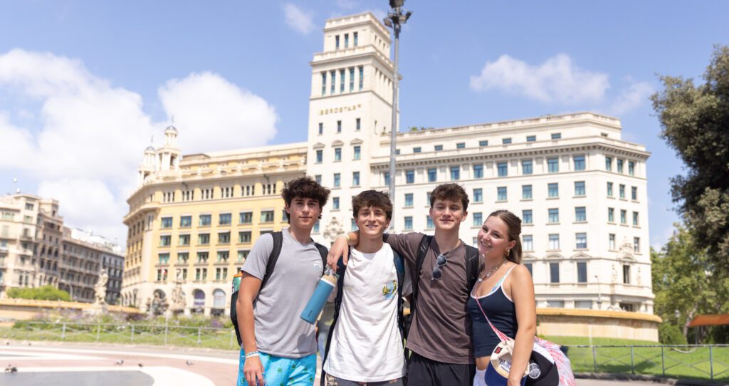 Four teens stand arm-in-arm smiling in front of a grand historic building in a city square on a sunny day, with blue skies and scattered clouds above.