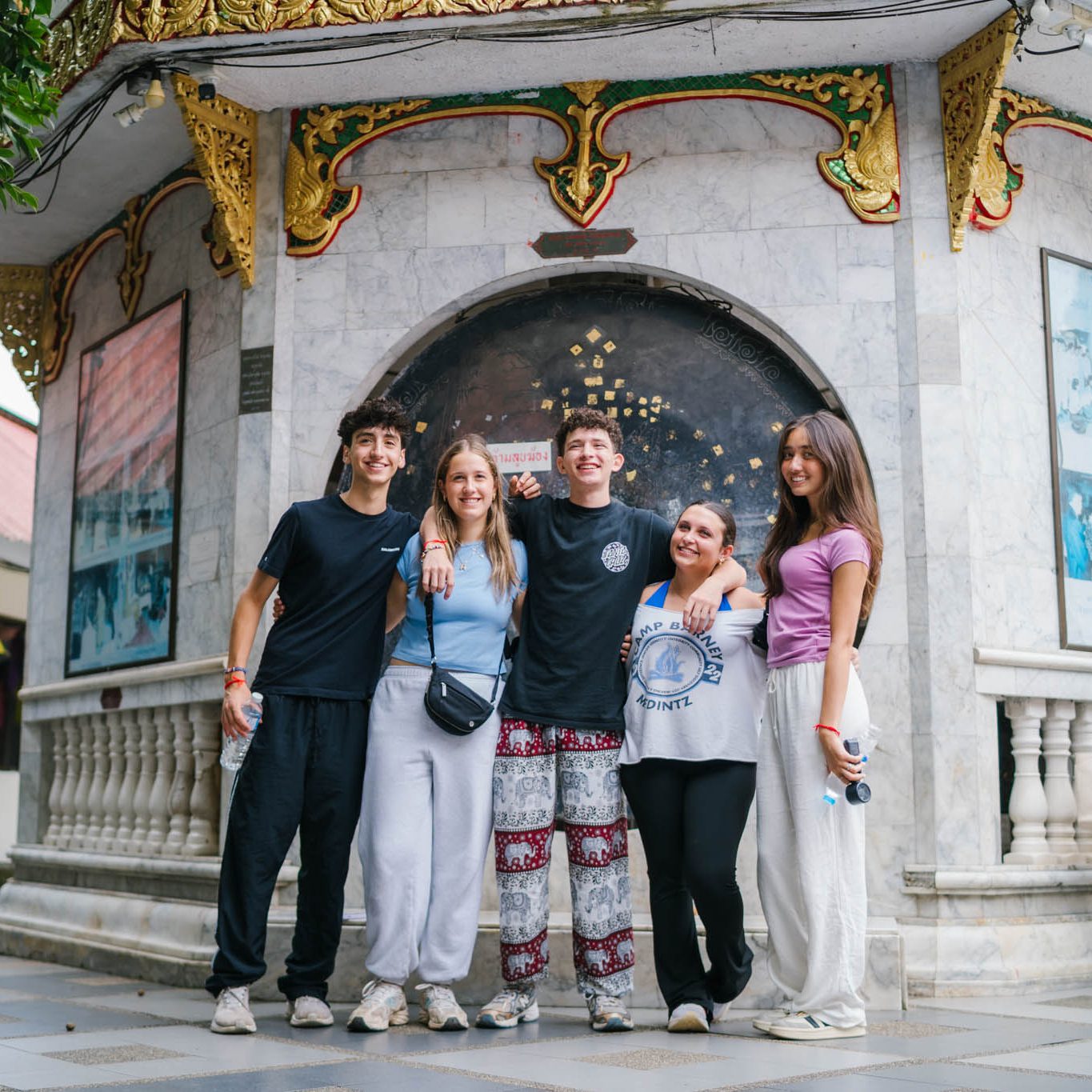 Ultimate-Thailand-Israel_Header-Photo_Square_DSC07167 Five teens stand arm-in-arm smiling in front of an ornate temple structure with gold decorations and marble walls, enjoying a sunny day.