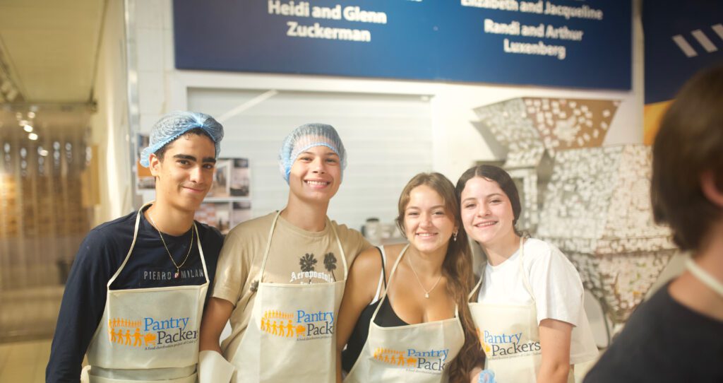 Four teens smile while volunteering at Pantry Packers, wearing aprons and some with hairnets, standing together inside the facility.