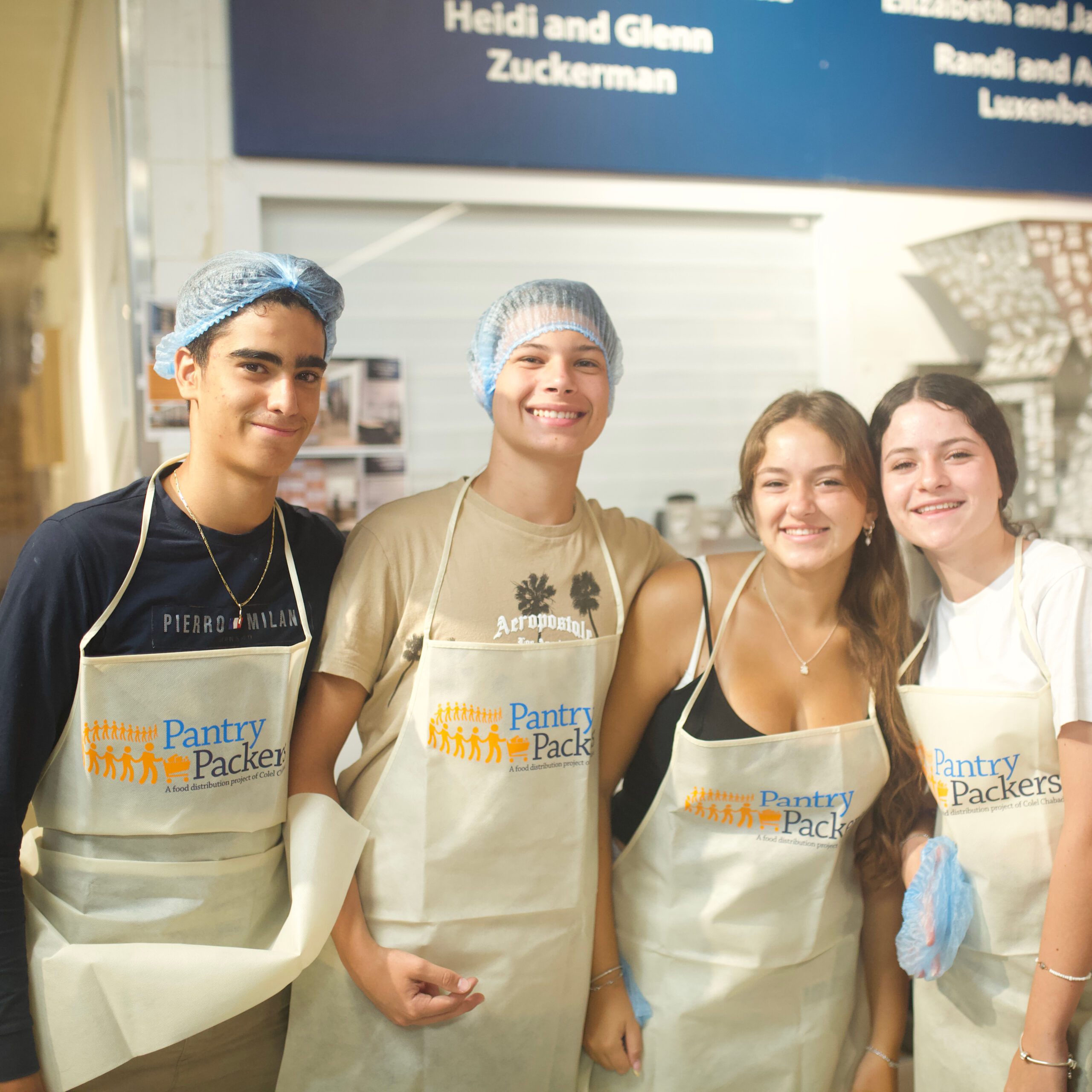 Volunteer-Israel_Header-Photo_Square_A34A2351 Four teens smile while volunteering at Pantry Packers, wearing aprons and some with hairnets, standing together inside the facility.